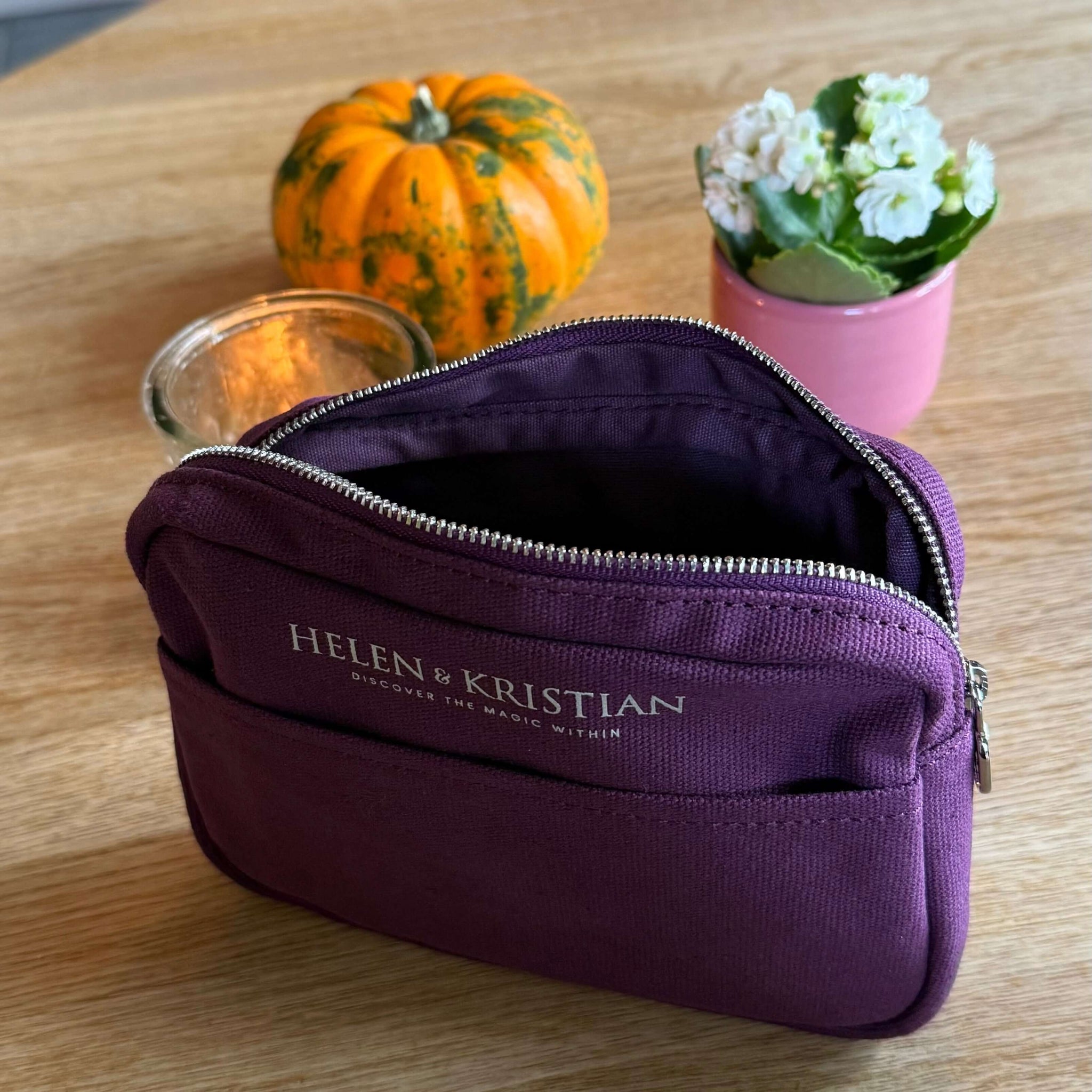 Purple cosmetic bag with 'Helen Kristian' branding on a wooden surface with a small pumpkin and plant in the background.
