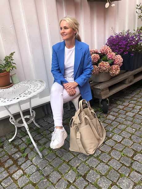 Woman in blue blazer and white outfit sitting at outdoor table with beige bag and flowers.